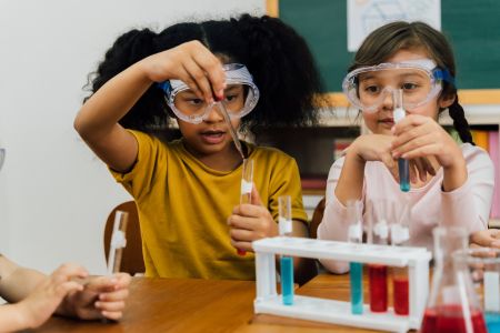 two young girls in a classroom wearing safety goggles doing an experiment 