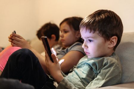 three children sitting on a couch side by side staring at electronic devices