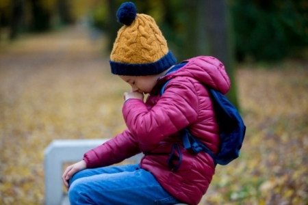 young boy in winter coat and hat sitting outside, coughing