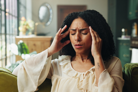 A woman sitting with her eyes closed and hands on her temples, indicating she has a headache and is stressed.